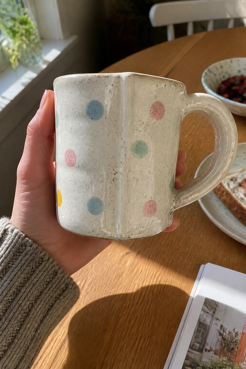 Grey-sleeved hand holding speckled ceramic mug with colorful polka dots