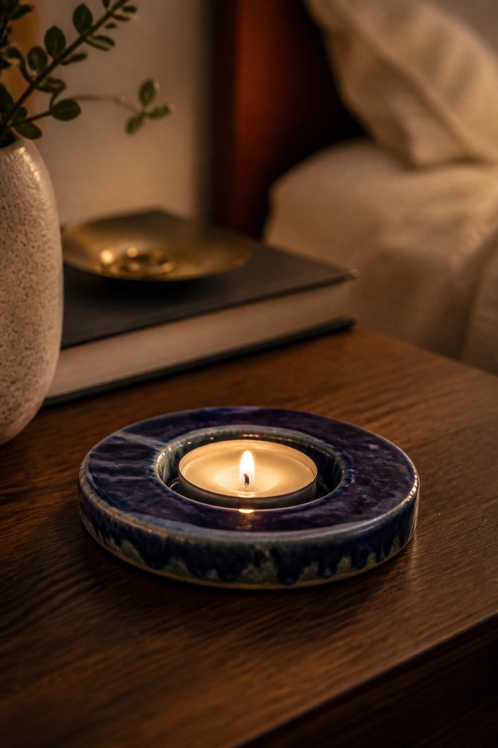 Lit candle in blue ceramic holder on wooden table with vase and books nearby.