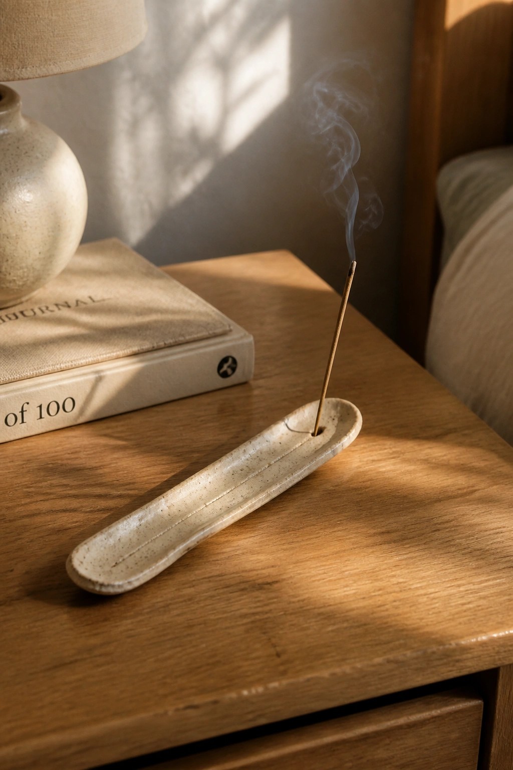 Burning incense in speckled ceramic holder on sunlit wooden nightstand beside journal