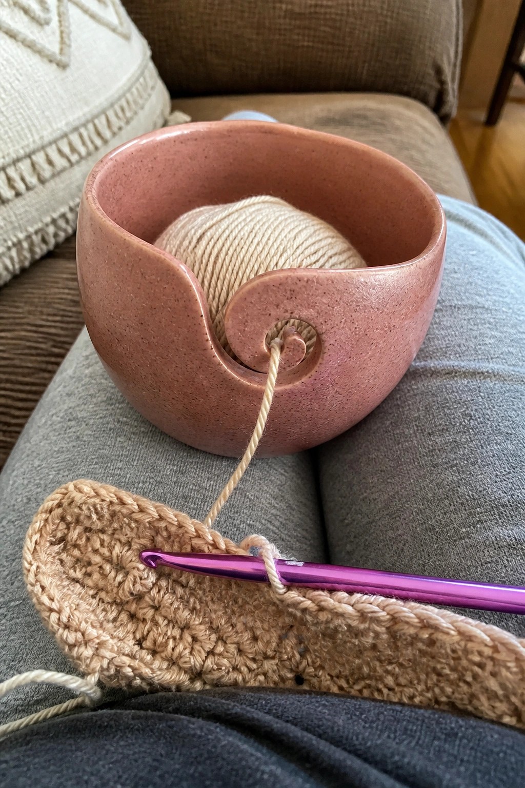 Pink ceramic yarn bowl with beige yarn, purple hook, and crochet project on lap