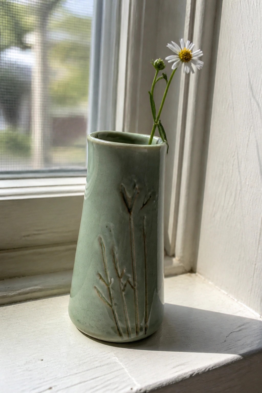 Green ceramic vase with reed etchings holding a white daisy on sunny windowsill.