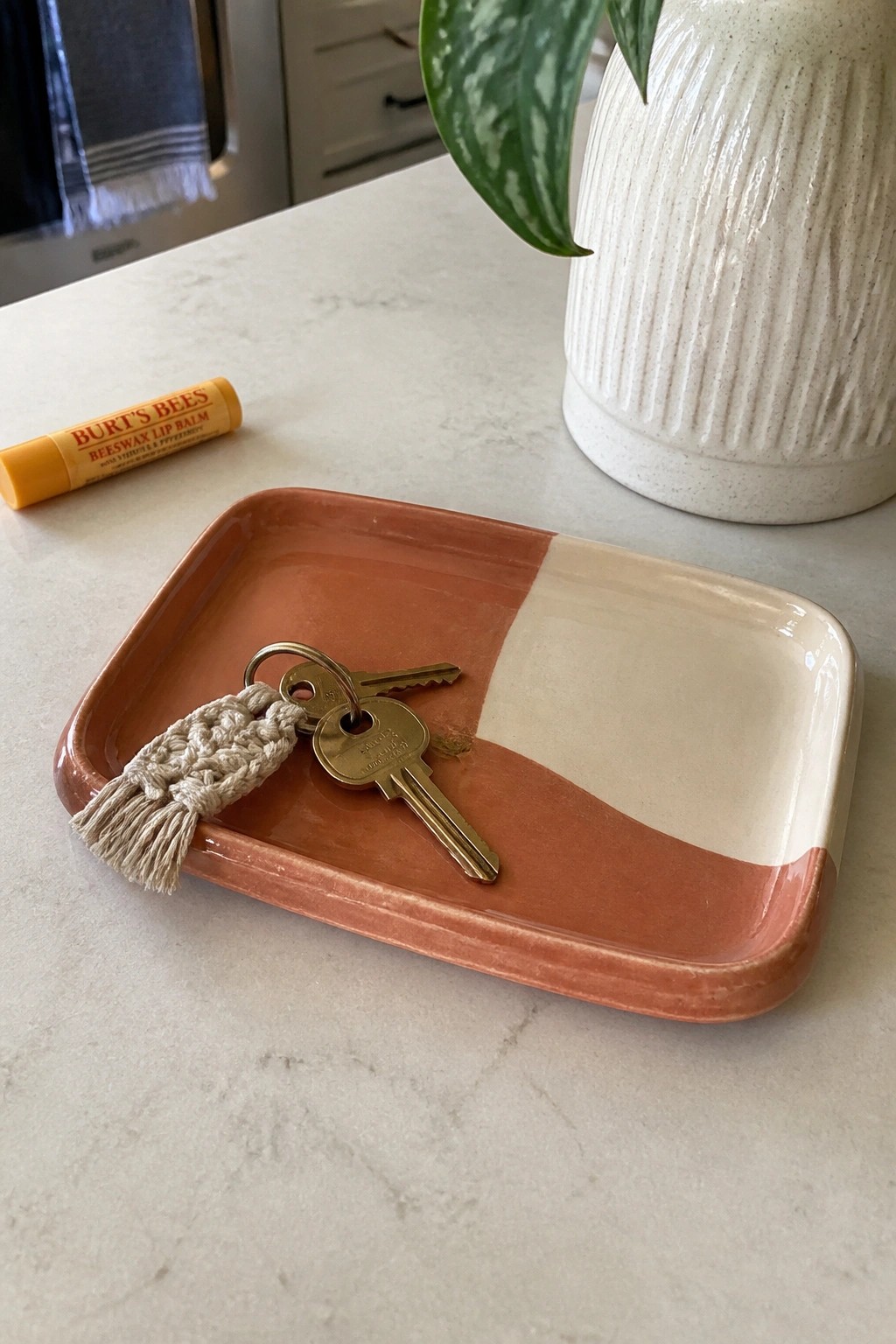 Keys with macrame tassel on two-toned ceramic tray beside lip balm on marble counter
