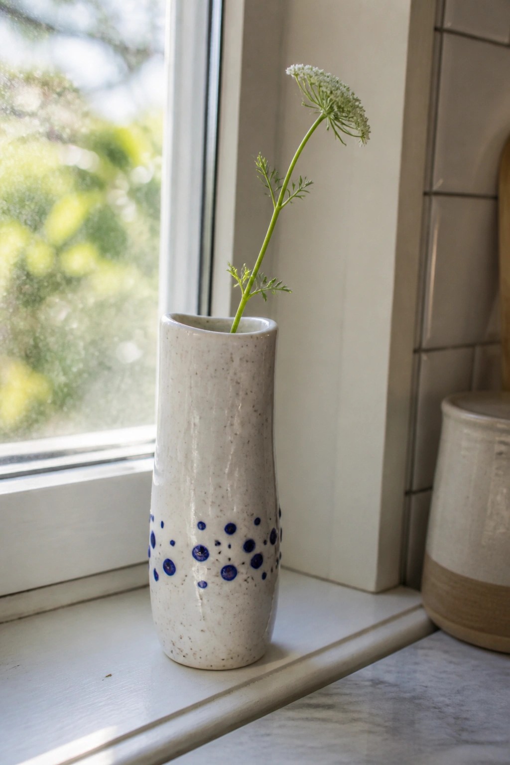 White ceramic vase with blue dots holding a green wildflower stem on windowsill