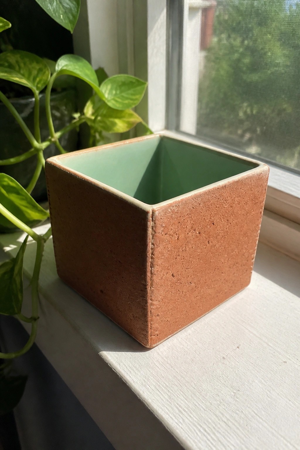 Square reddish-brown ceramic pot with green interior on white windowsill near plant