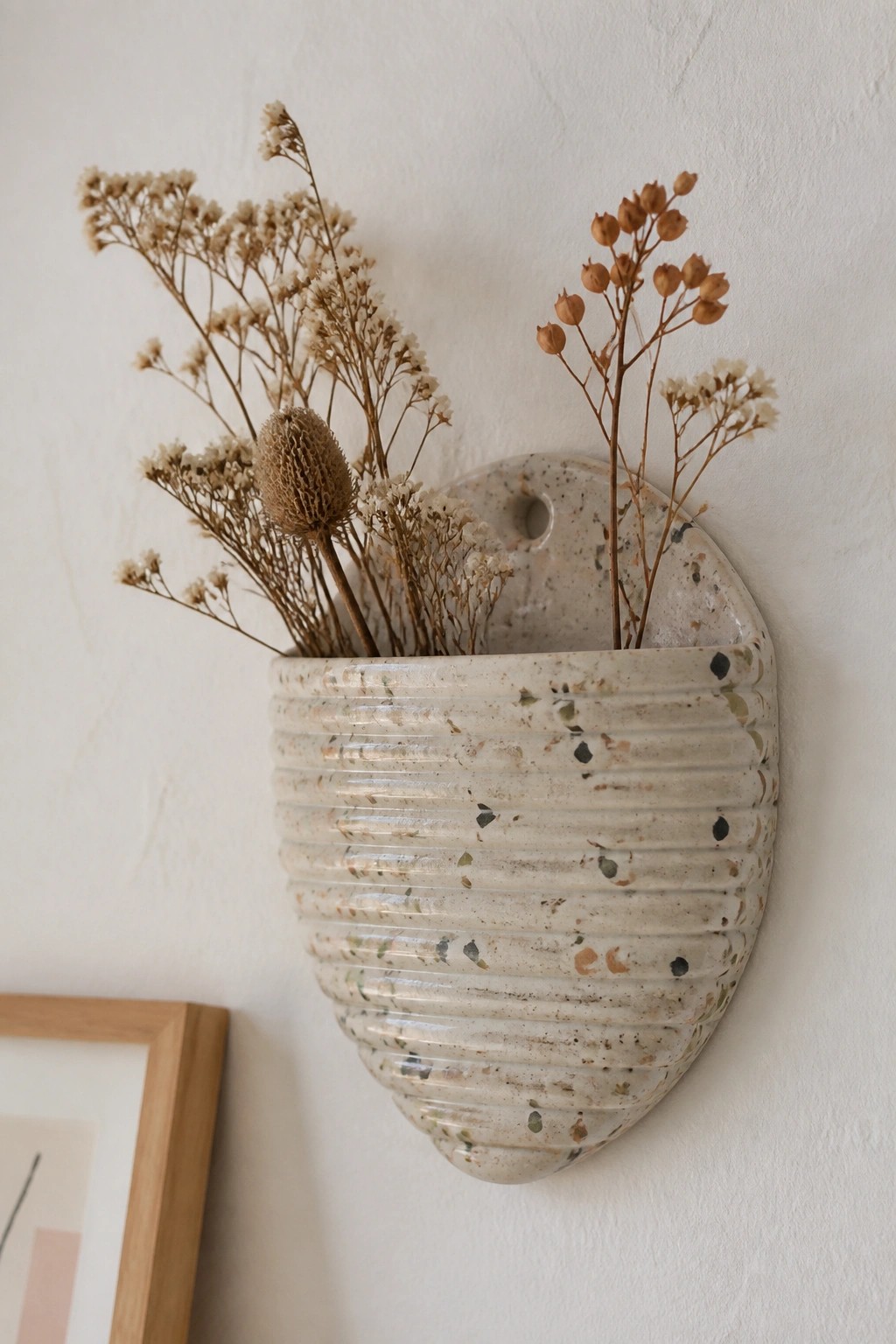 Speckled beige ribbed ceramic wall vase holding dried flowers on white wall