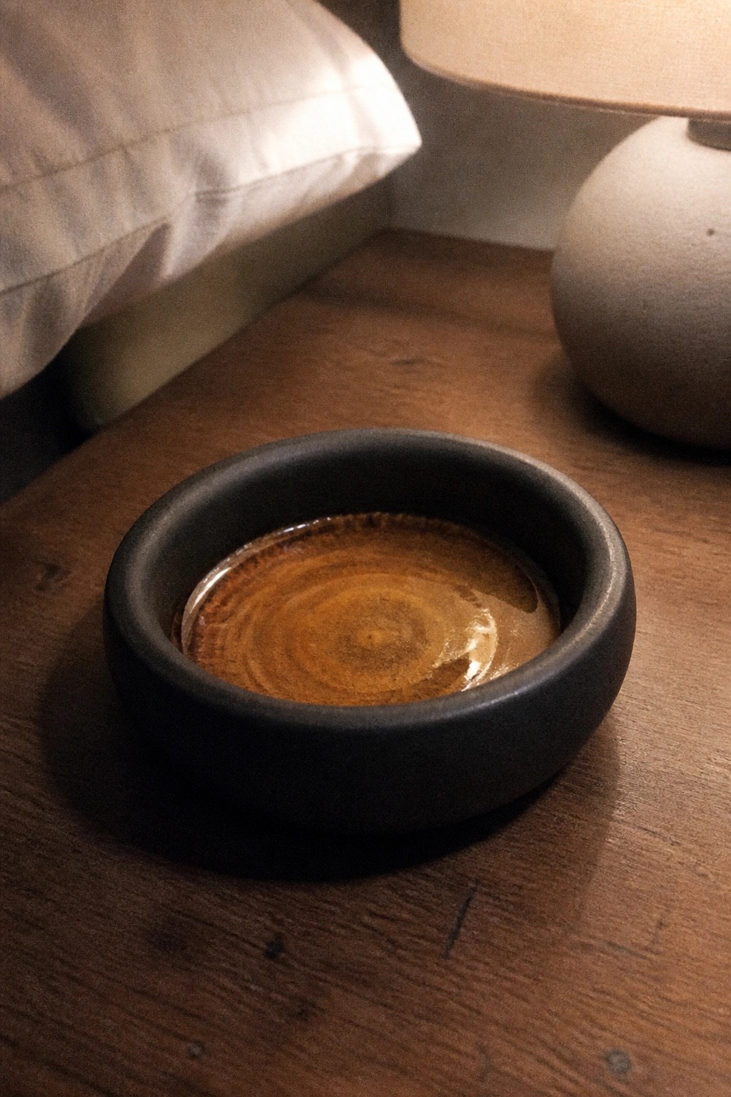 Black bowl of swirling brown coffee on wooden table near lamp and pillow