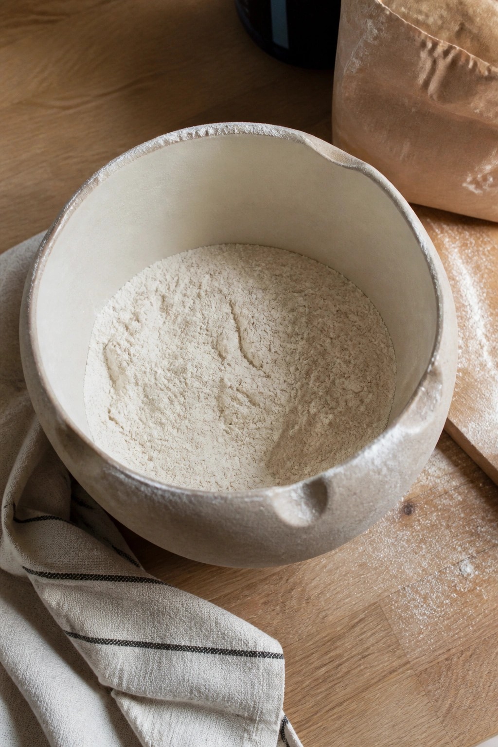 Ceramic bowl of flour on striped linen atop wooden table with paper bag