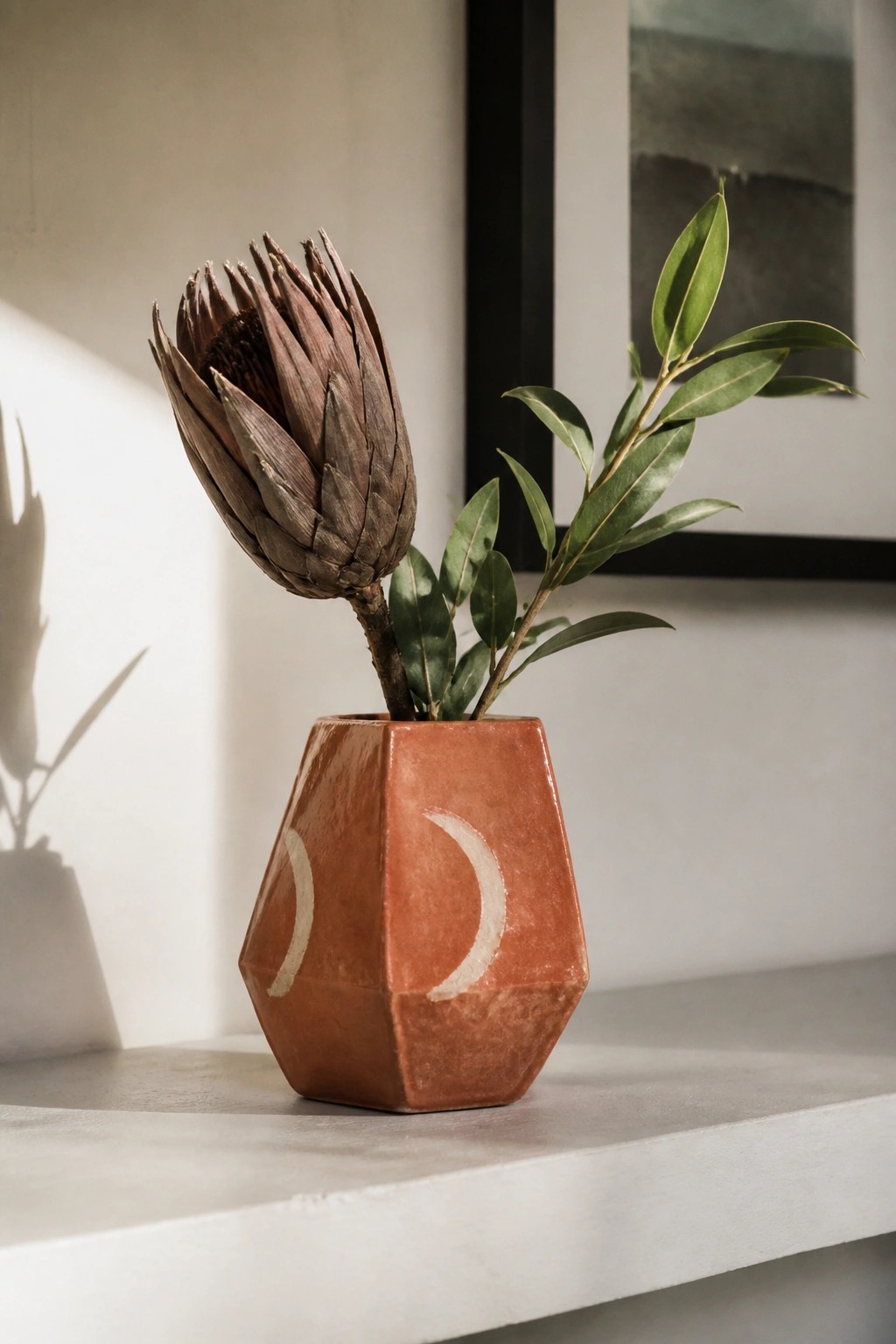Faceted terracotta vase with white crescents holding dried protea and green leaves
