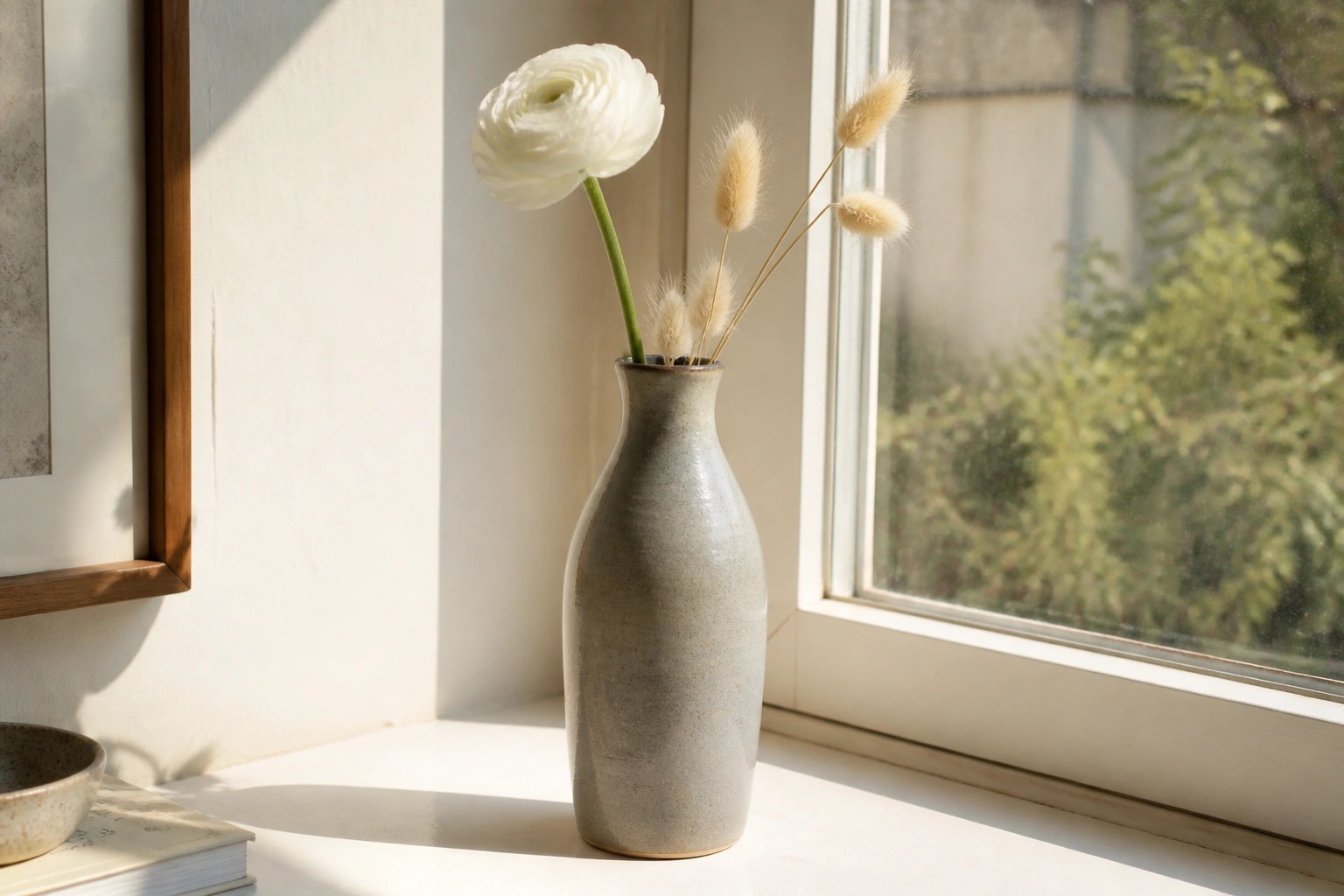 White ranunculus and dried grasses in a speckled vase on a sunny windowsill