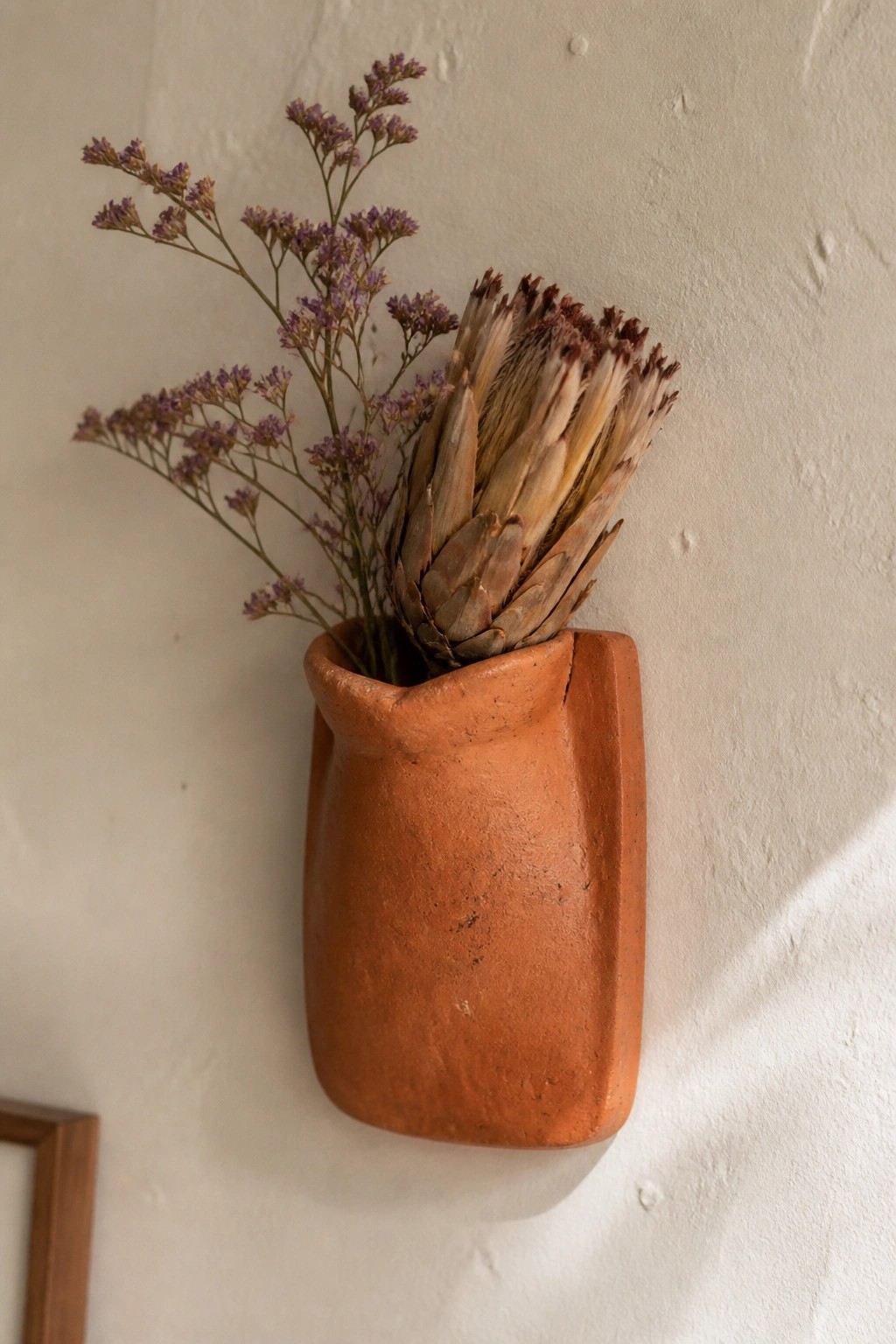 Terracotta wall pocket vase holding dried protea and purple flowers on white wall
