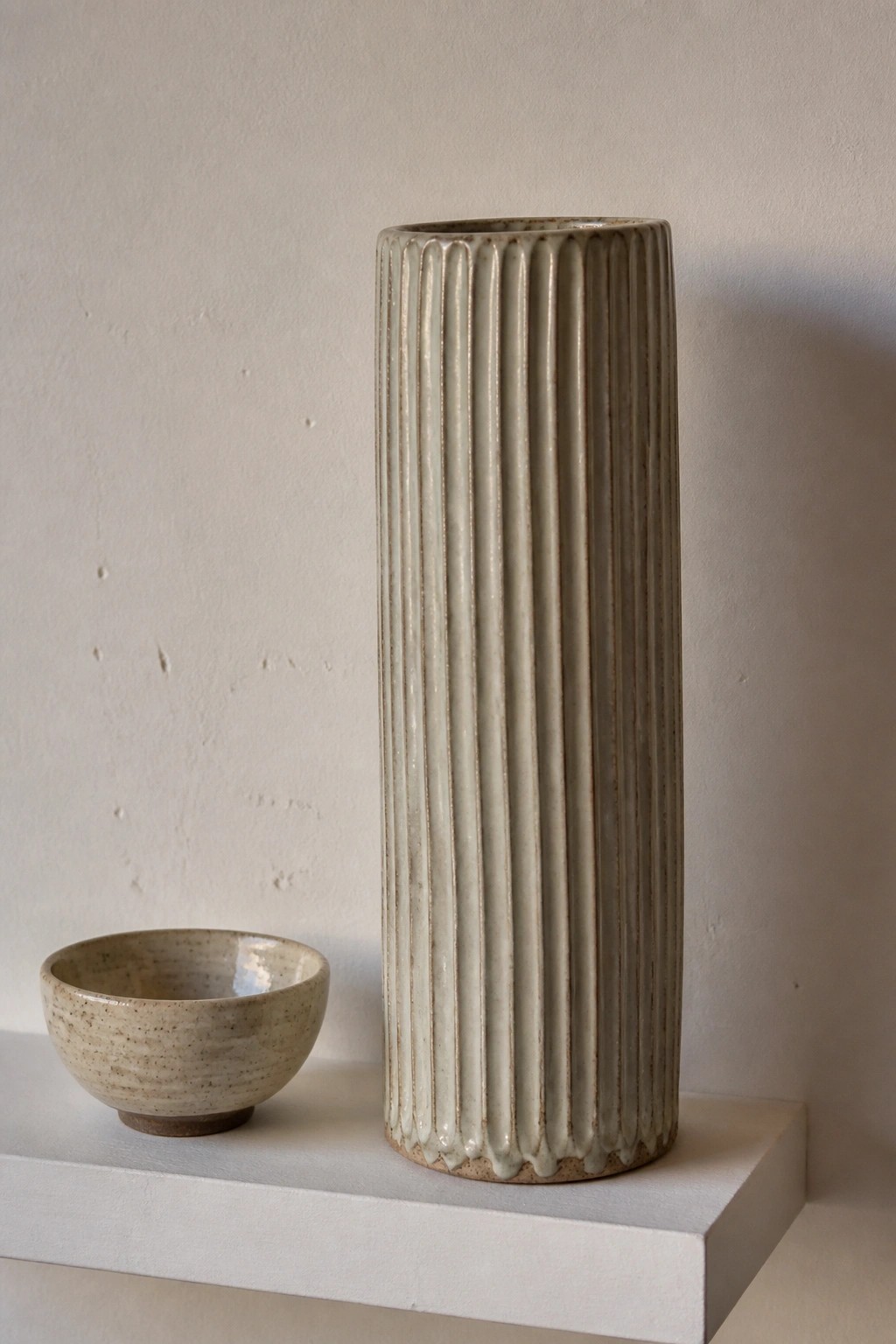 Tall ribbed ceramic vase beside small speckled bowl on white shelf.