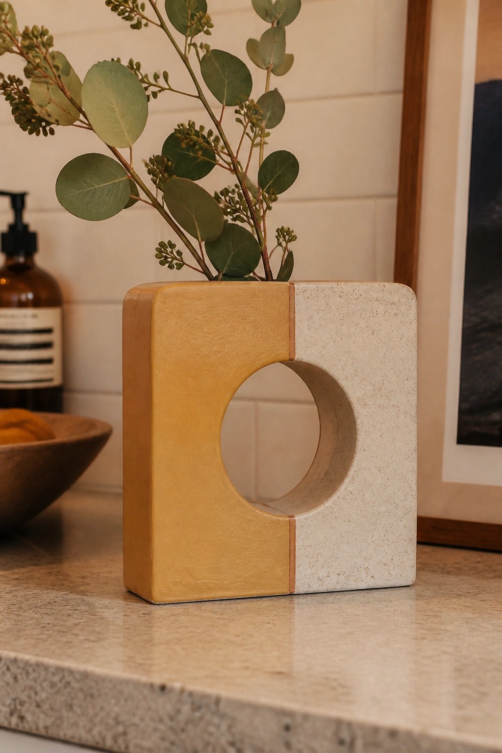Square two-tone yellow and white vase with circular cutout holding eucalyptus on marble counter