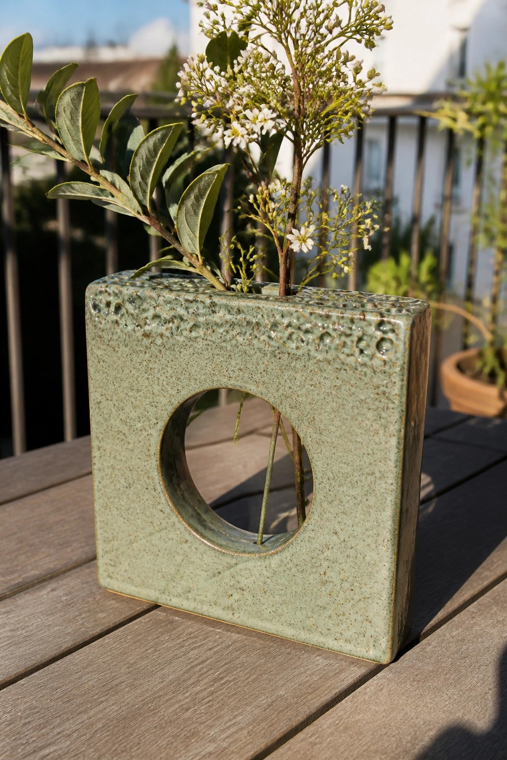 Green square ceramic vase with circular hole holding white flowers on wood