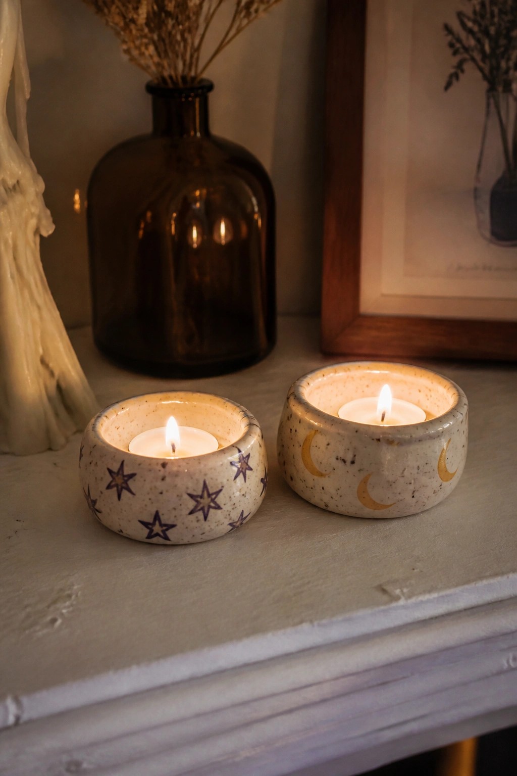 Two small ceramic bowls painted with stars and moons hold lit tea light candles on a light wooden surface.