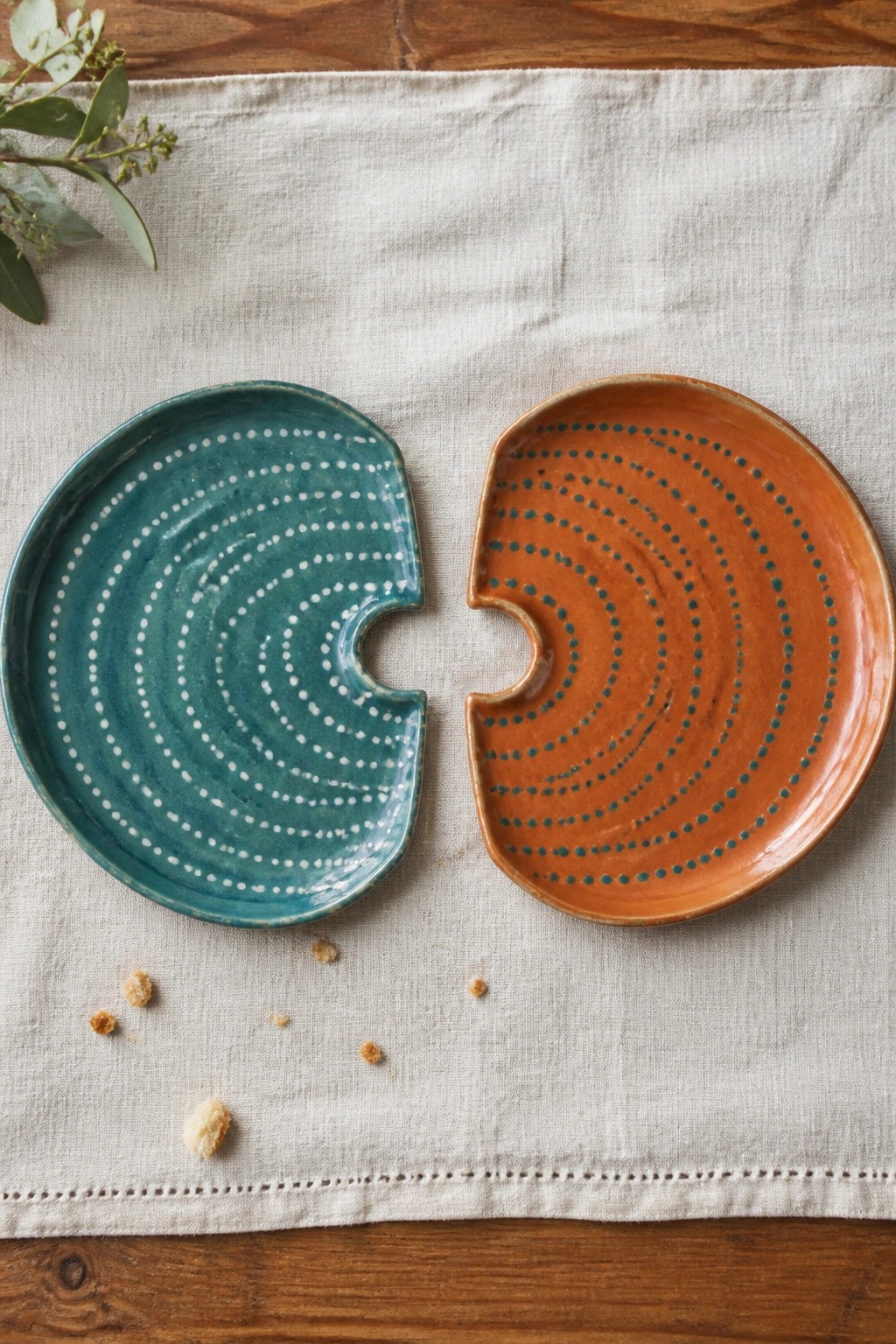 Two interlocking ceramic plates, teal with white dots and orange with teal dots, on linen.