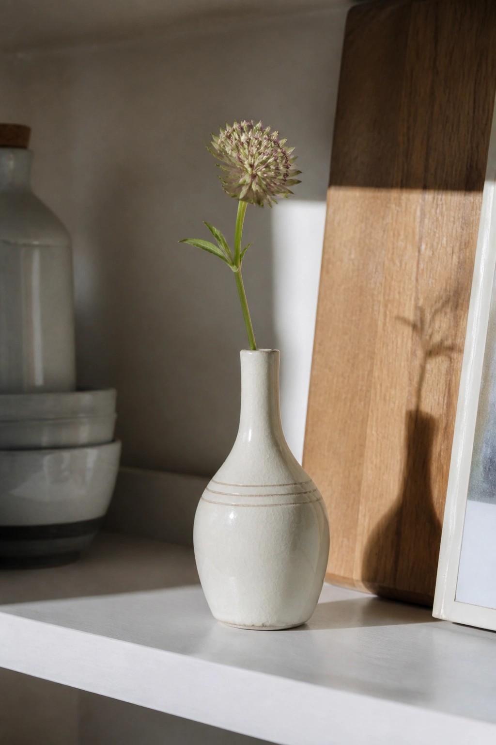 White ceramic vase holding one spiky green-purple flower on a shelf