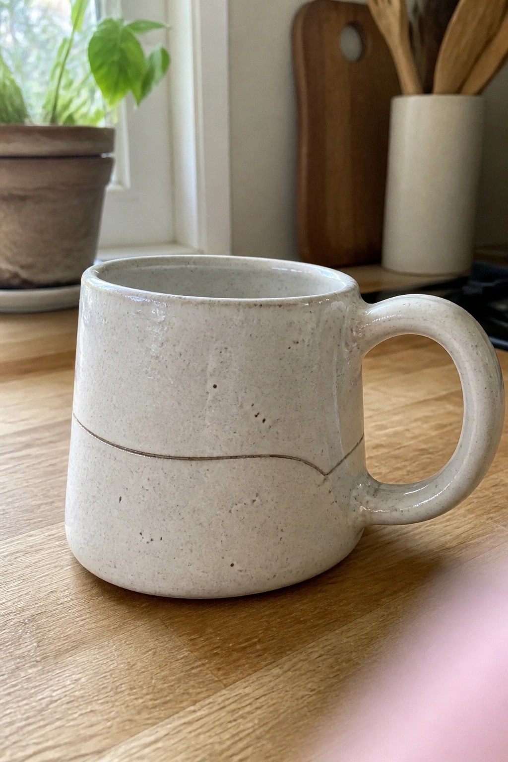 A short off-white speckled ceramic mug with one thin wavy incised line around the body rests on a wooden surface.
