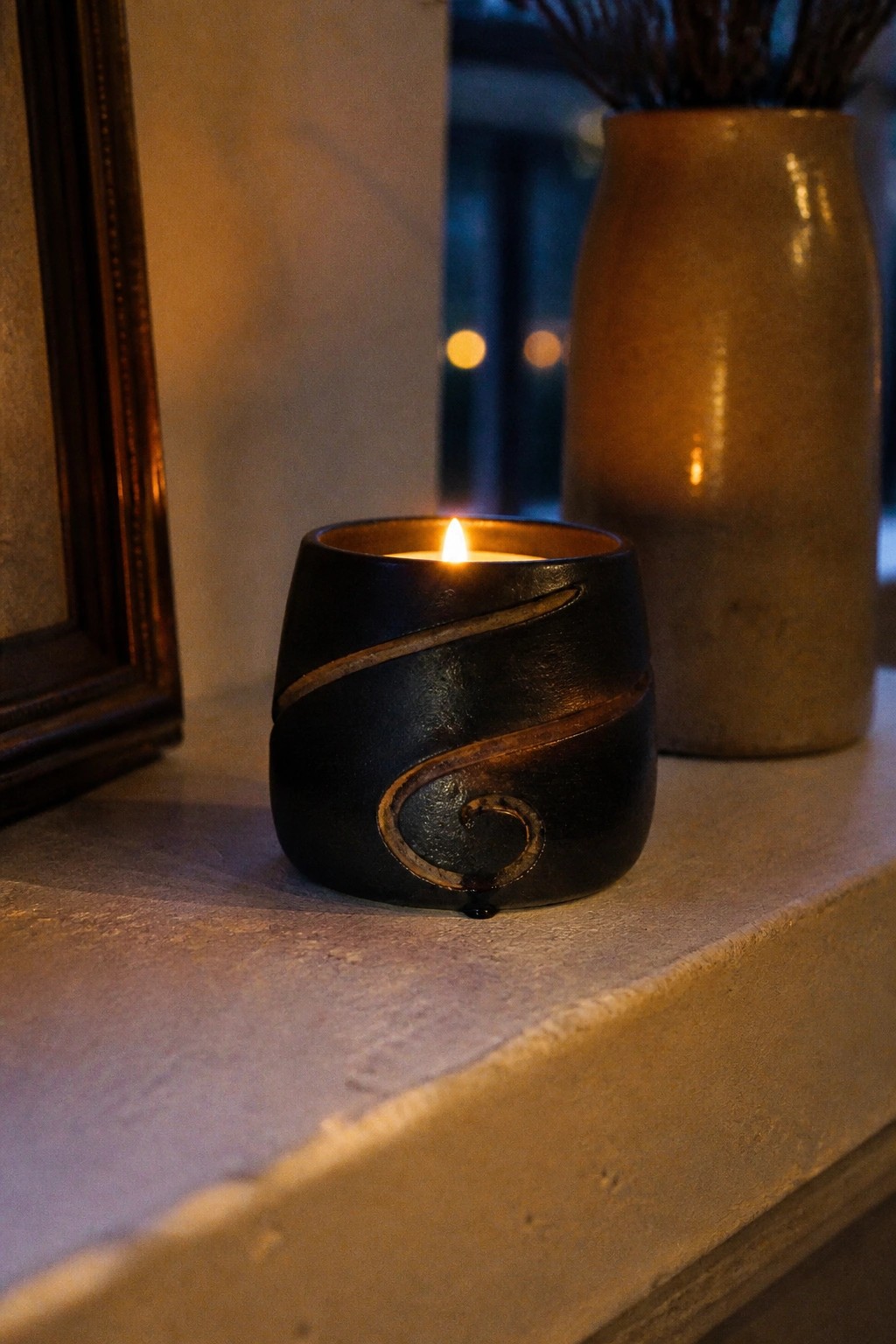 Lit candle in black ceramic holder with gold swirl on stone ledge beside vase