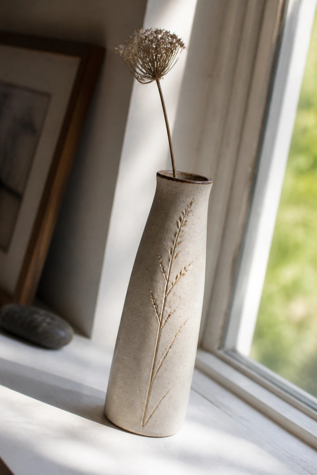 Cream-colored ceramic vase with raised grass design holding dried flower on windowsill