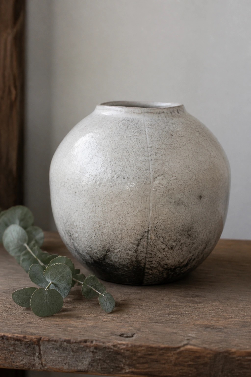 Round crackled ceramic vase beside eucalyptus leaves on rustic wooden table