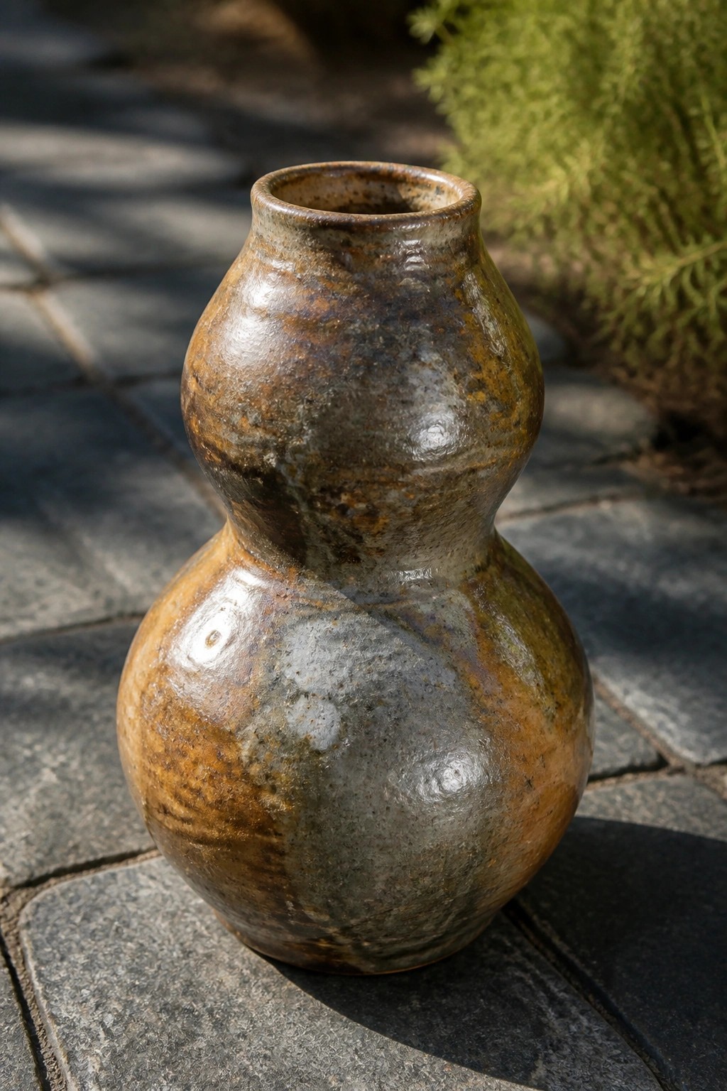 Rustic ceramic double-gourd vase with mottled brown, gray, and green glaze on stone.