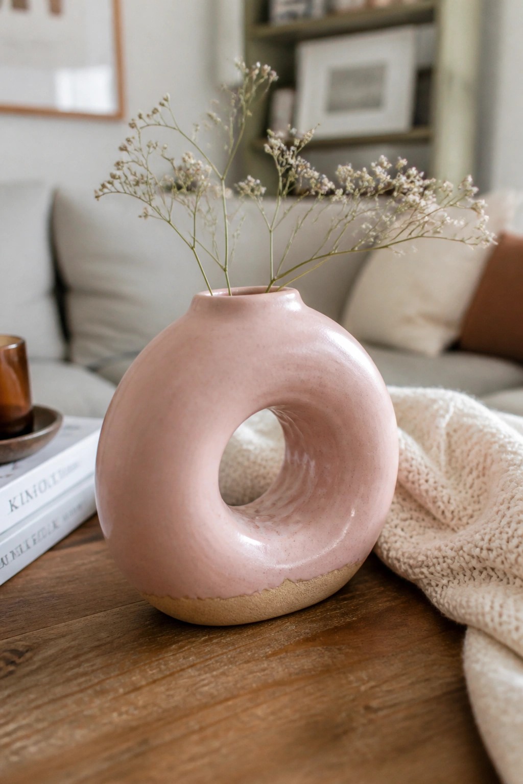 Pink donut-shaped ceramic vase with dried flowers on wooden table beside knit blanket