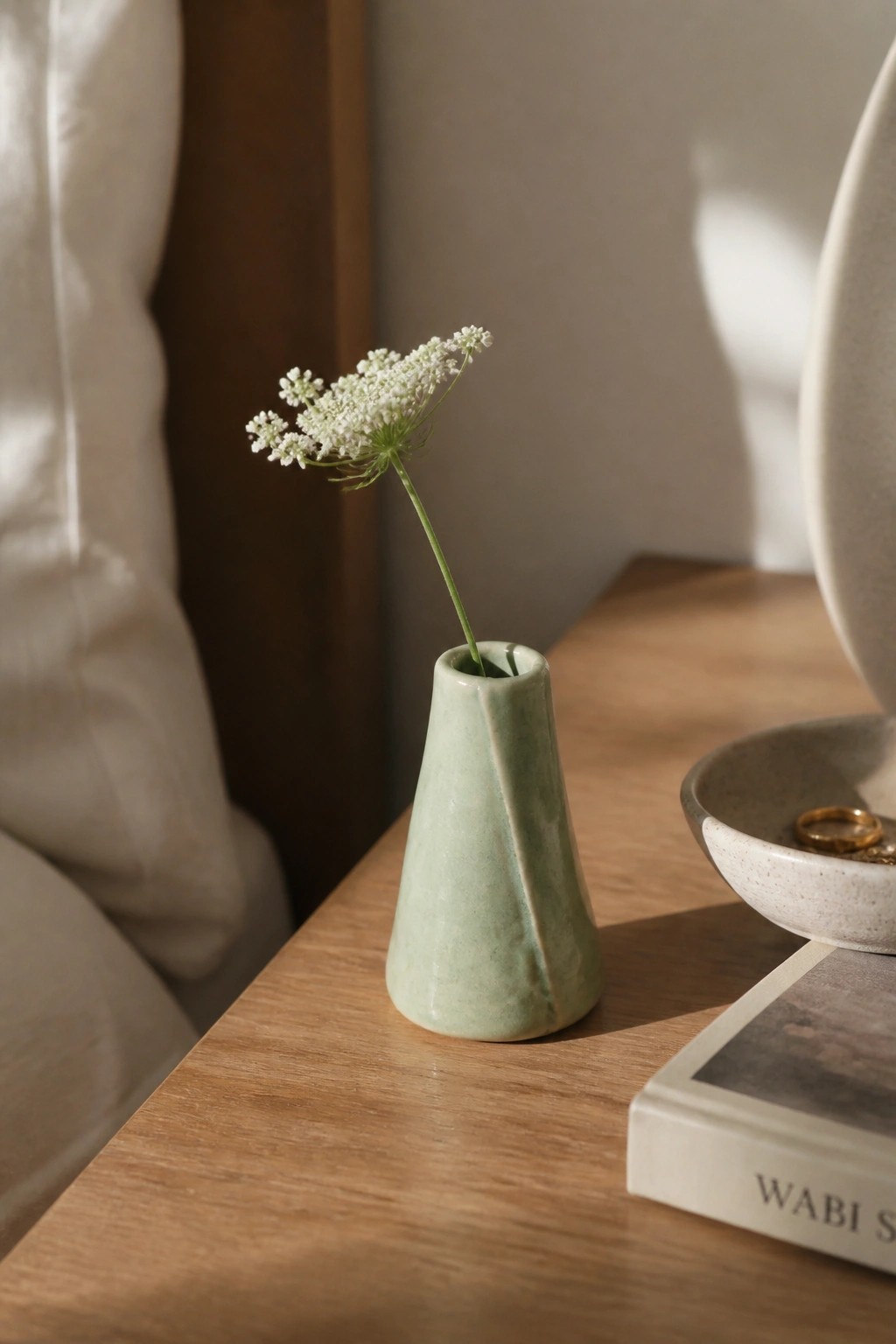Light green ceramic vase with white flower on wooden table beside book and bowl.