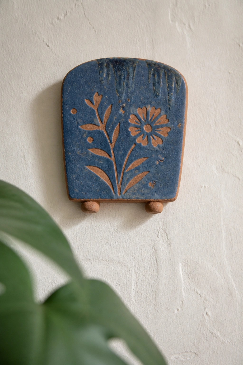 Blue ceramic tile with brown floral etching on white wall near green leaf