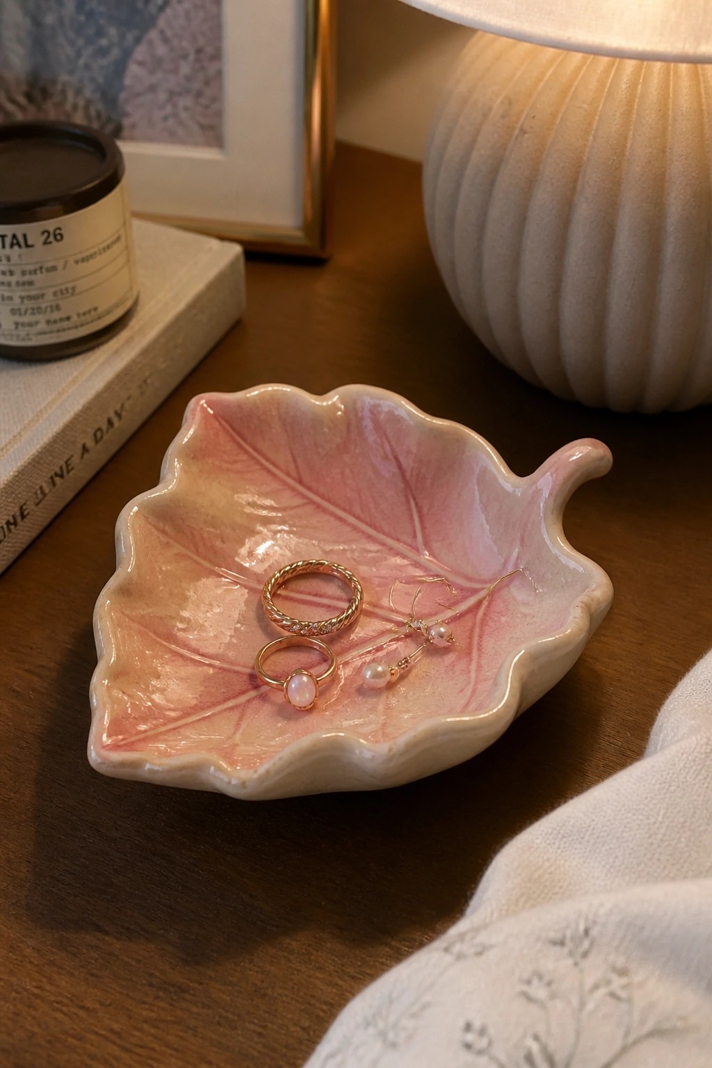 Pink ceramic leaf dish holding gold rings and pearl earrings on wood.