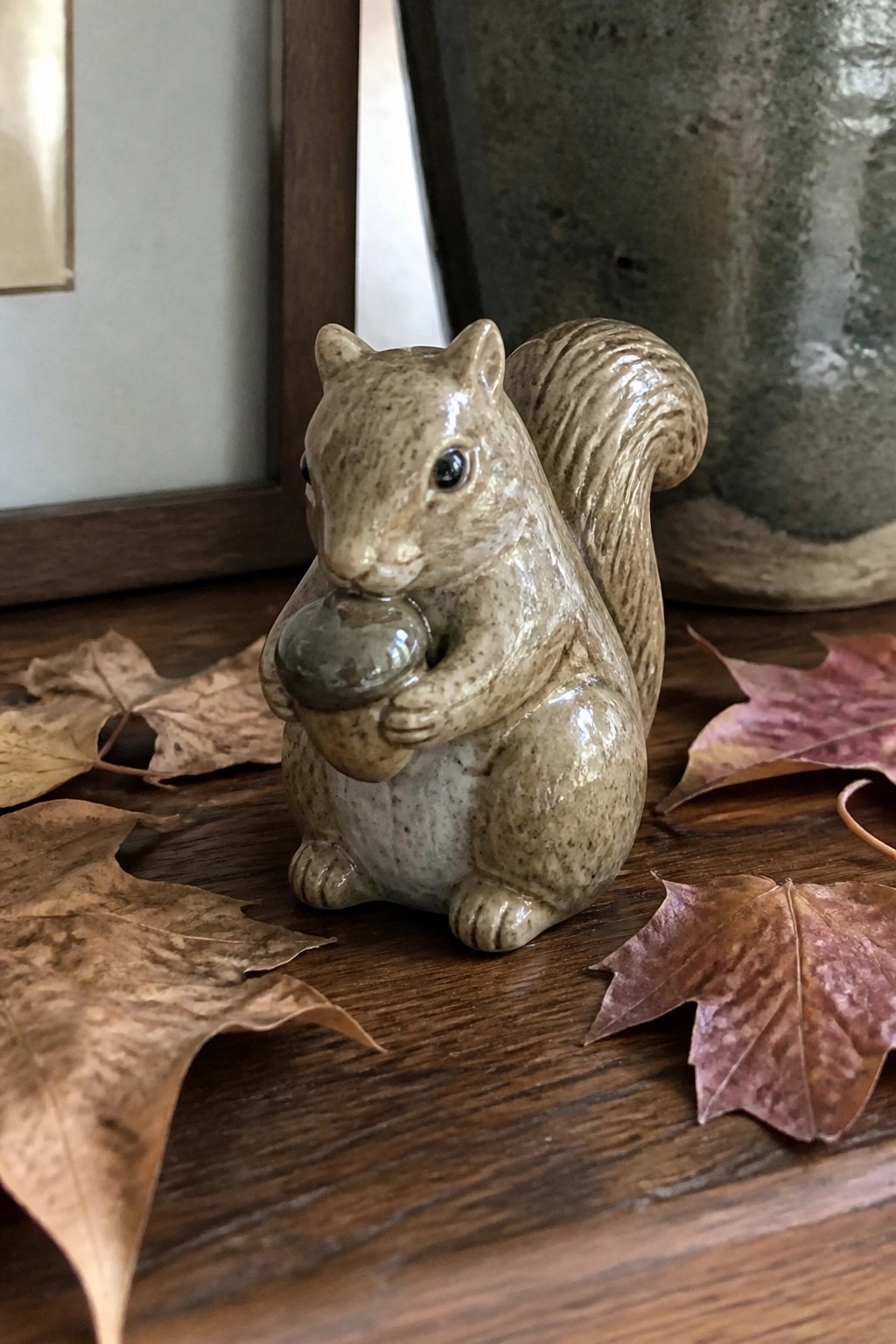 Ceramic squirrel figurine holding acorn amid dried autumn leaves on wooden table