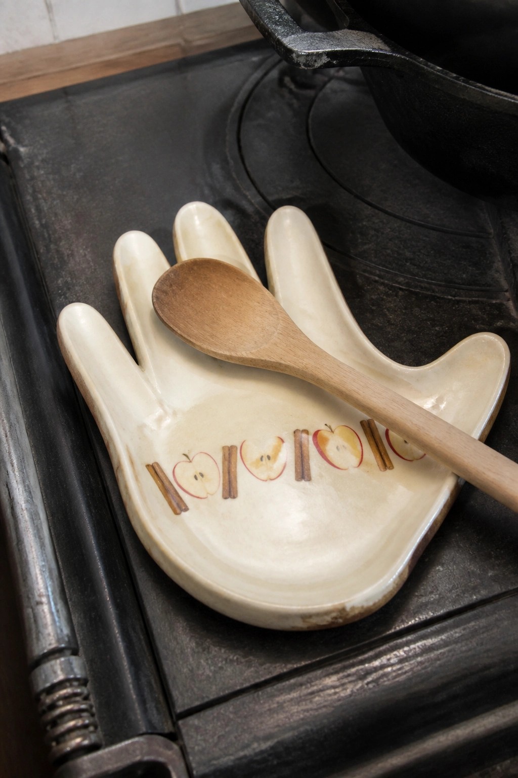 A ceramic hand-shaped dish with painted apples and cinnamon sticks holds a wooden spoon on a black cast iron stove.