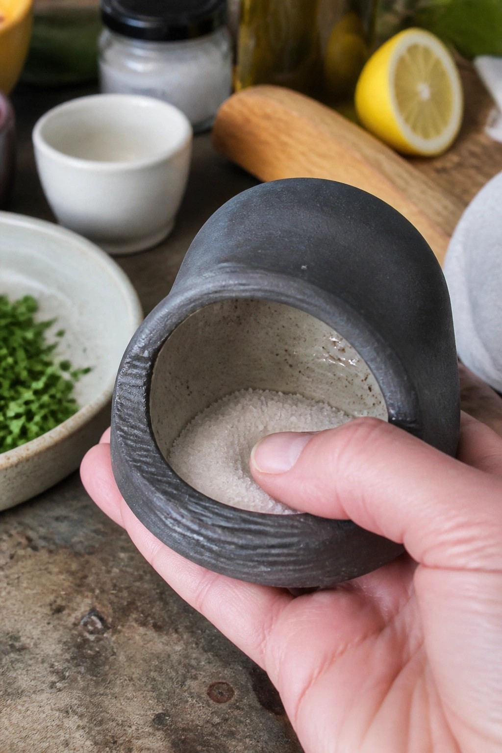 Hand holding dark ceramic bowl of white salt, thumb touching grains
