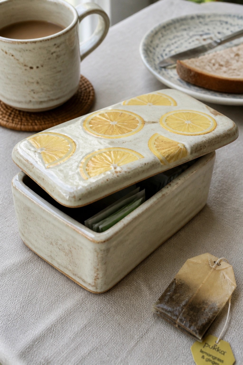 Open ceramic tea box with lemon slices beside used pukka teabag, mug, and toast plate