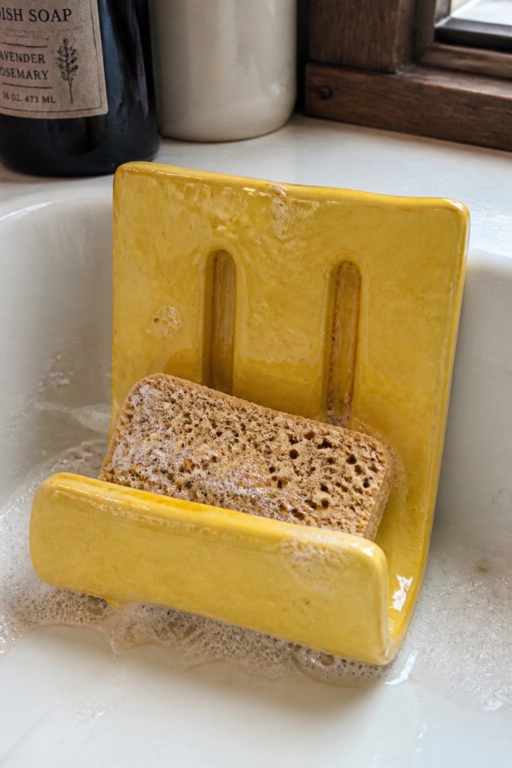 Yellow ceramic holder cradling wet brown sponge in bubbly white sink