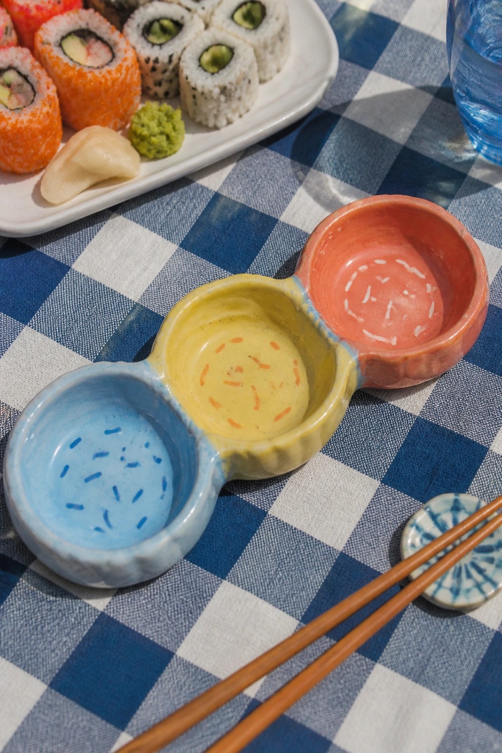 Colorful triple ceramic sauce bowls beside sushi on blue checkered tablecloth