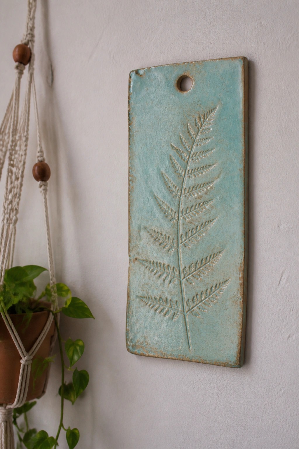 Turquoise ceramic plaque with embossed fern hanging on white wall beside macrame plant.