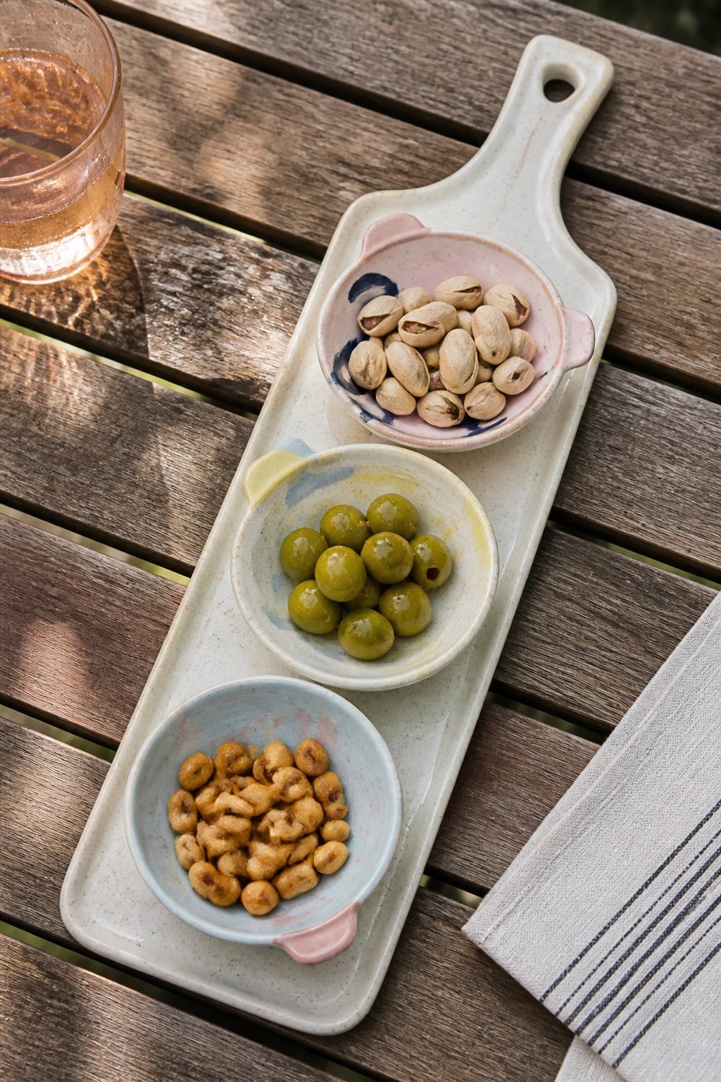 Three bowls of pistachios, olives, and nuts on tray with pink drink on wood table.