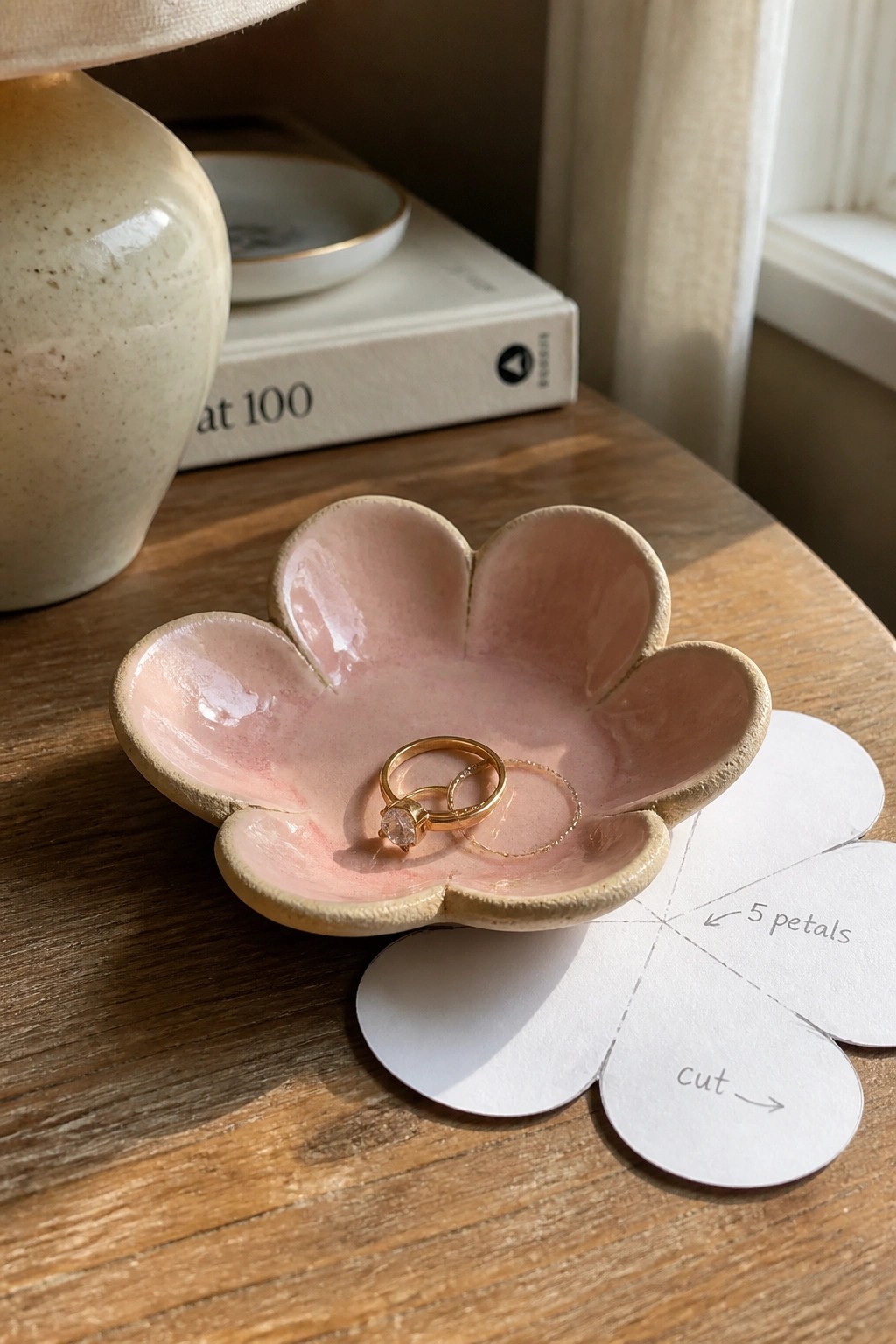 Pink flower-shaped ceramic bowl with gold rings beside paper 5-petal cutout on wood table