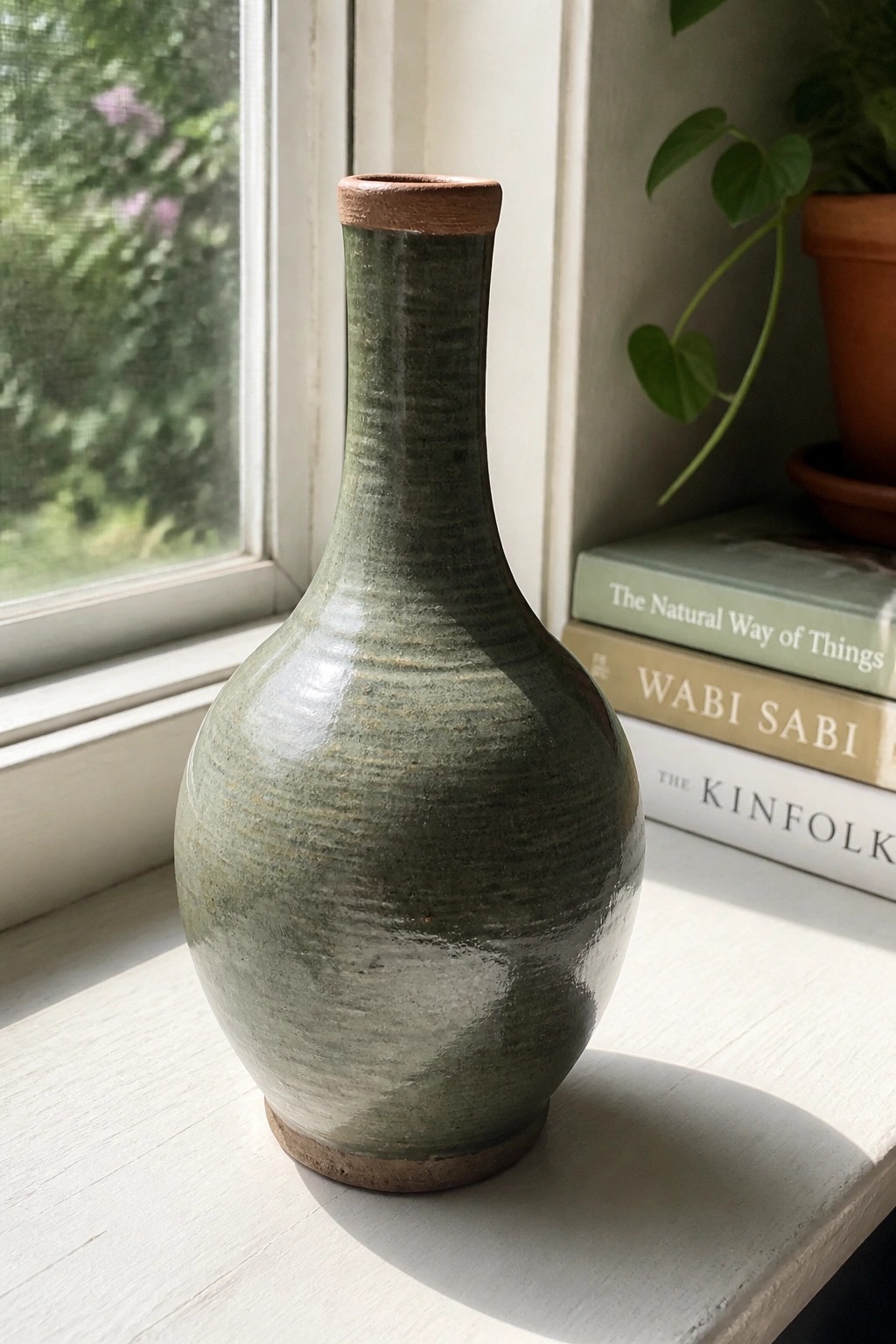 Green ceramic vase on sunny windowsill beside stacked books and houseplant.