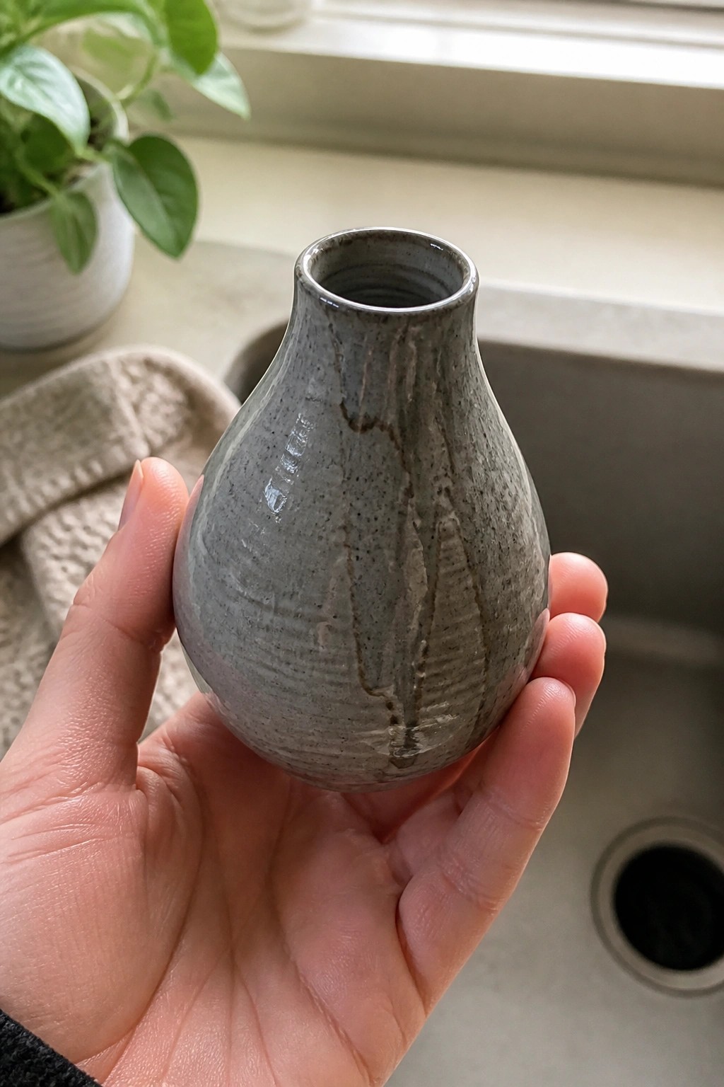 Hand holding a small gray ceramic vase above a kitchen sink with plant nearby