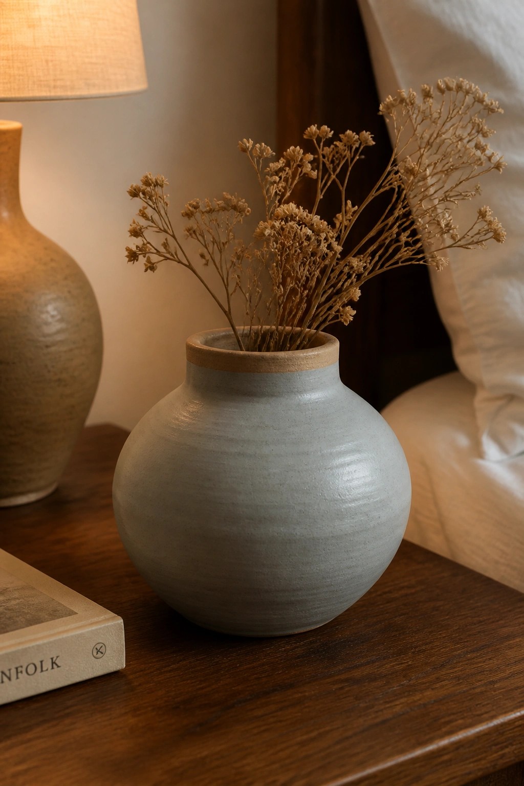 Gray ceramic vase with dried flowers on wooden table beside lamp and book