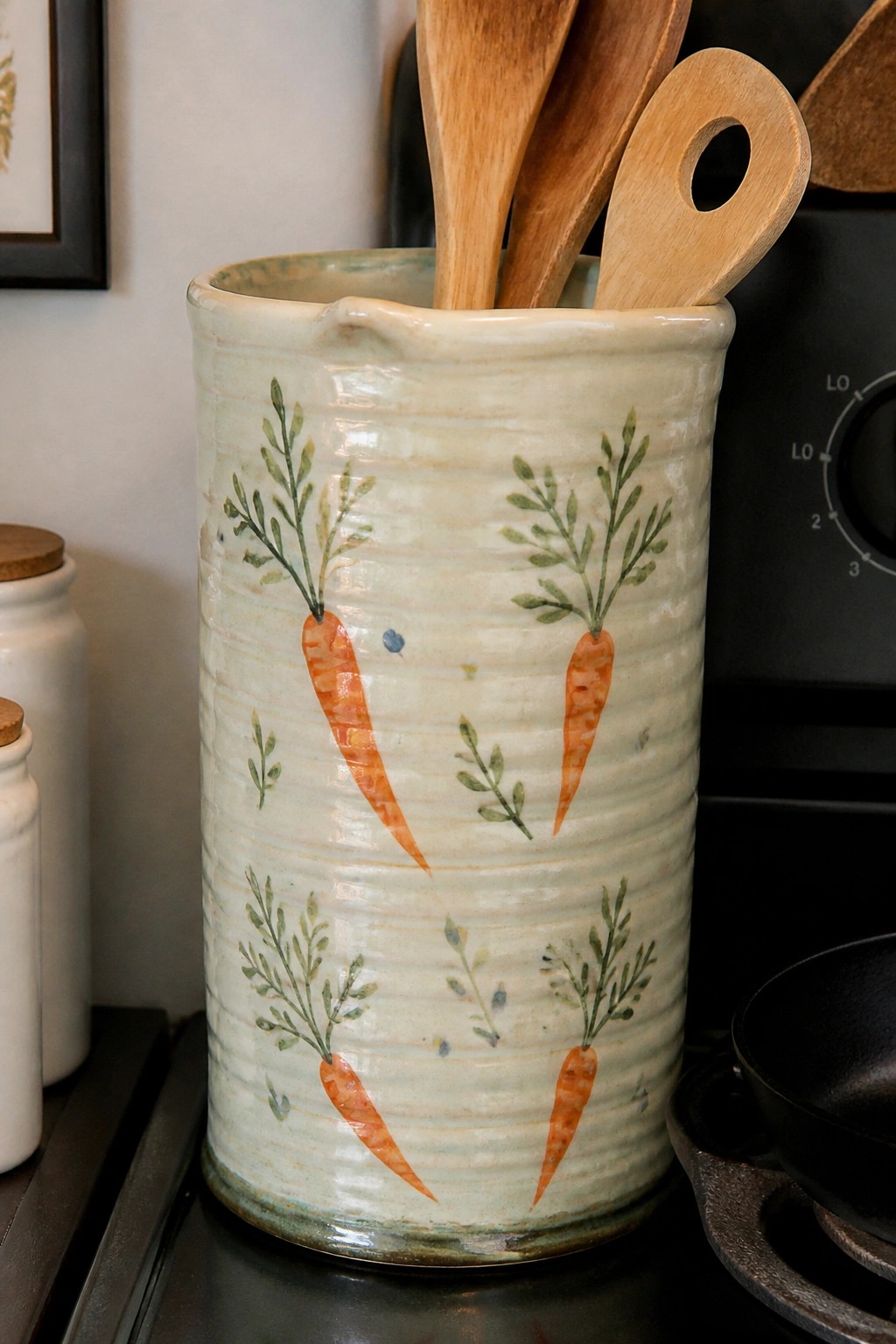 Ceramic crock painted with orange carrots holding wooden kitchen utensils on stove