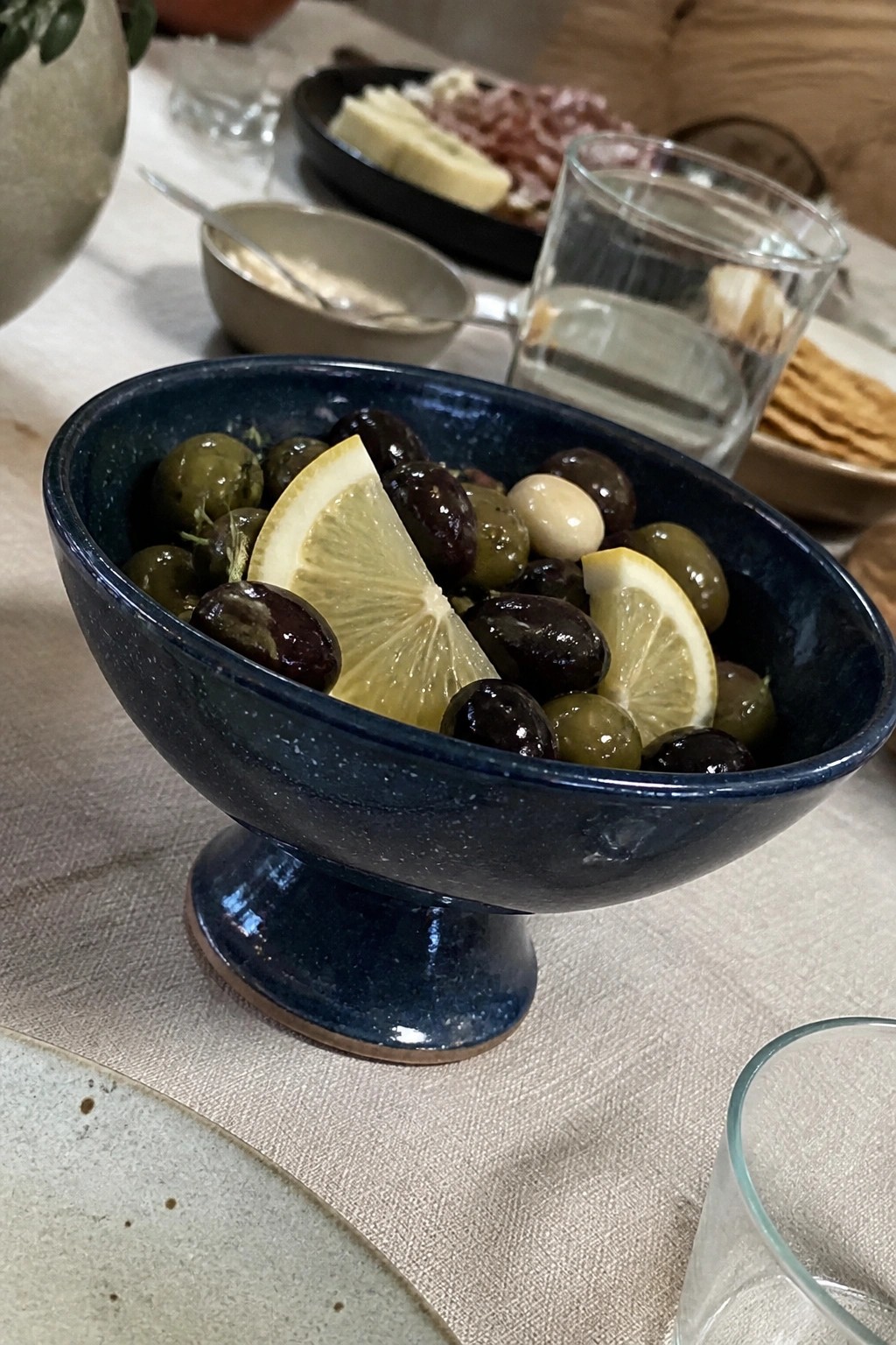 Blue ceramic pedestal bowl of mixed olives with lemon slices on table