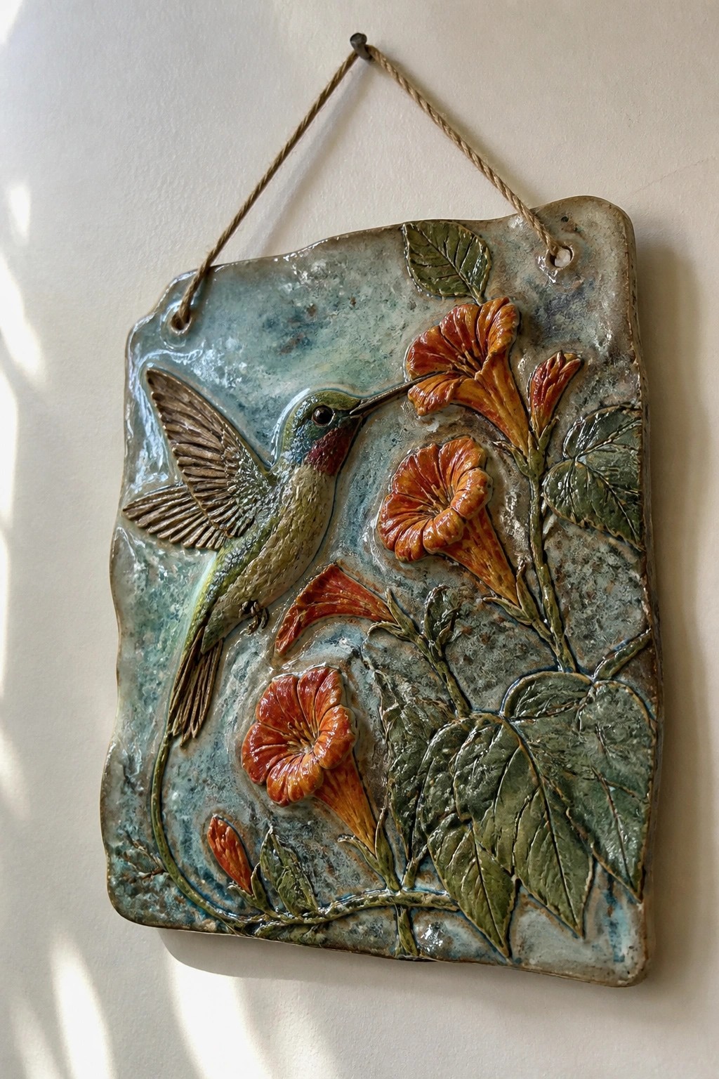 Ceramic relief plaque of a hummingbird amid orange trumpet flowers and green leaves.