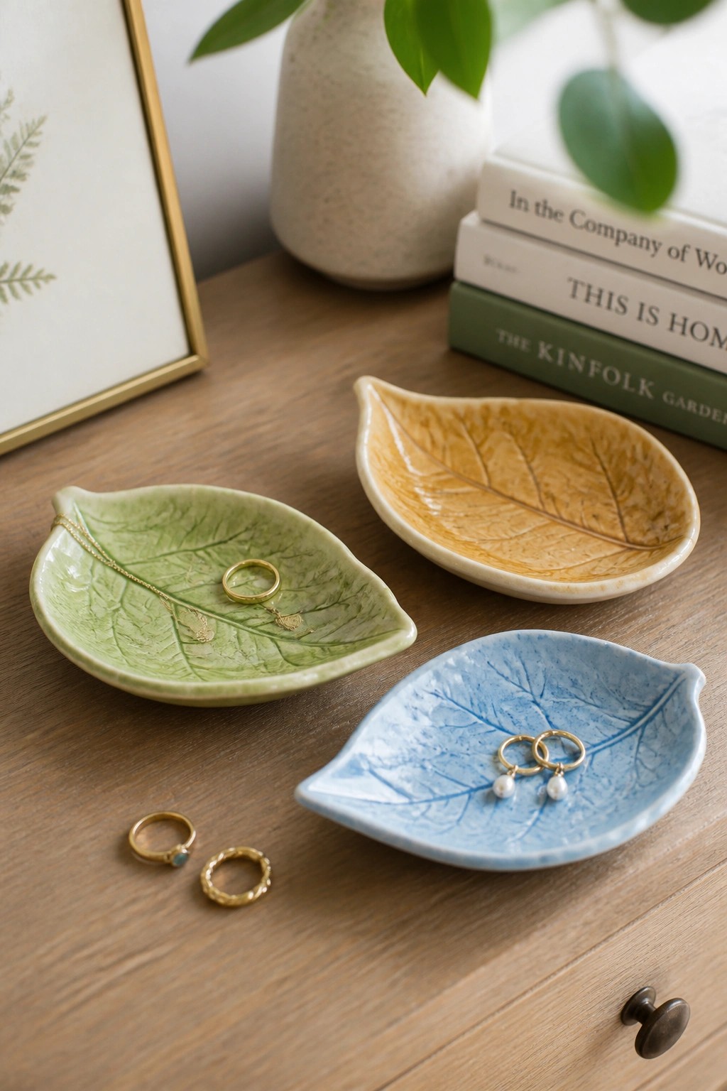 Three ceramic leaf bowls in green, yellow, and blue holding gold jewelry on wood