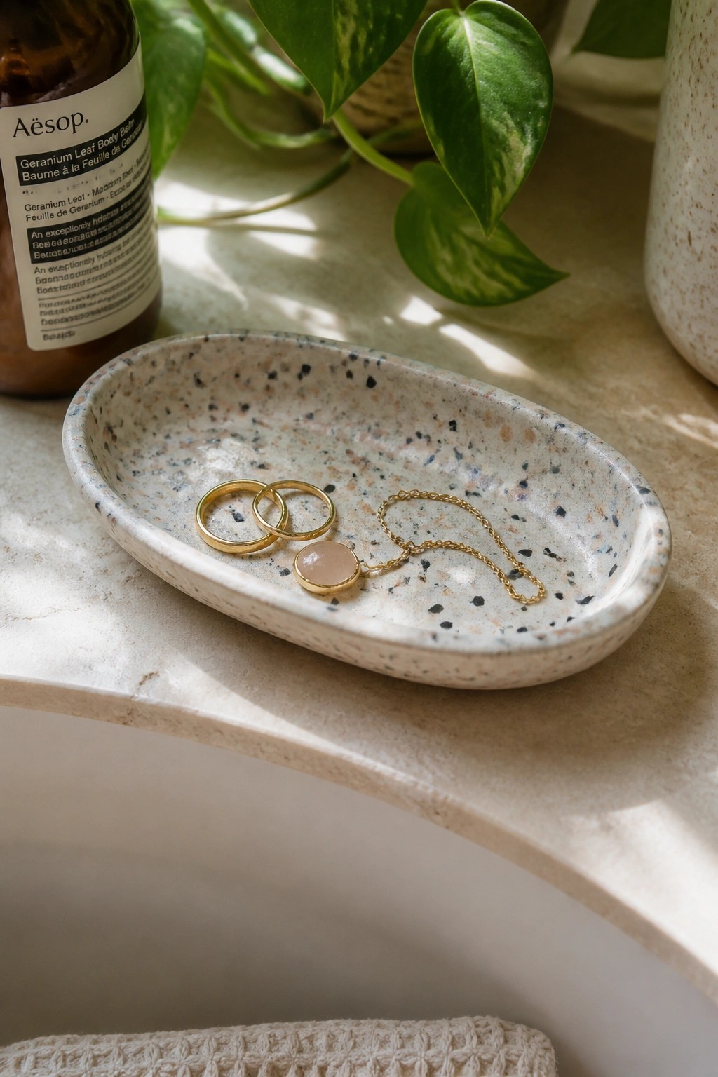 Oval speckled tray holding two gold rings and pink pendant necklace on marble.