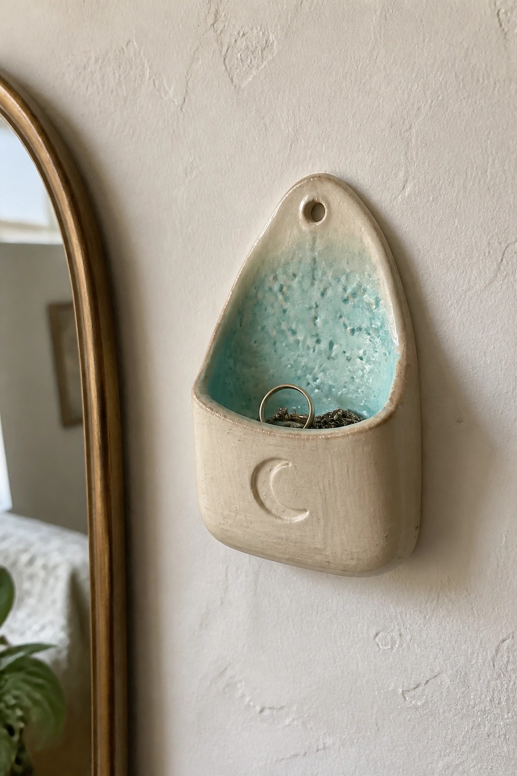Ceramic wall pocket with turquoise interior holding rings beside gold mirror frame
