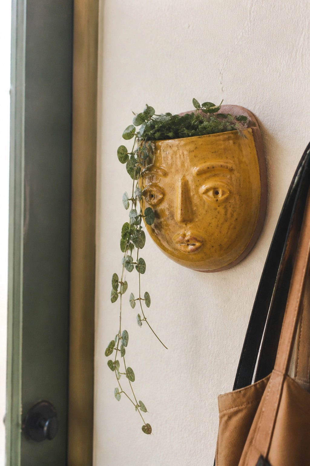 Yellow ceramic face planter with trailing vines on white wall beside brown bag