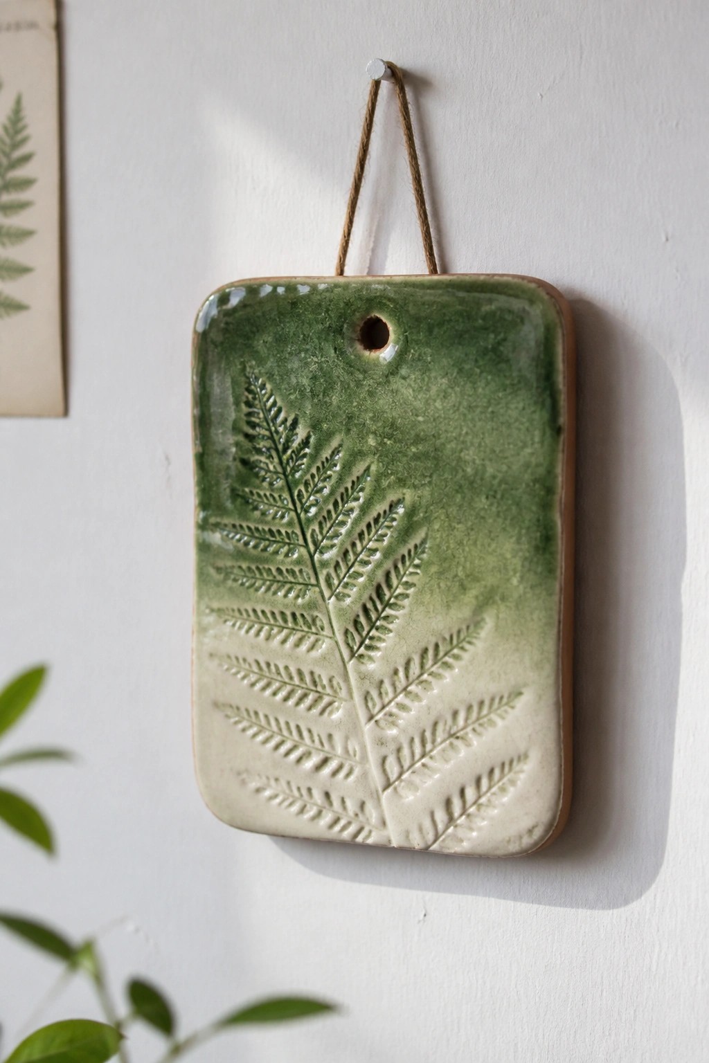 Green ceramic plaque with embossed fern design hanging on white wall by twine