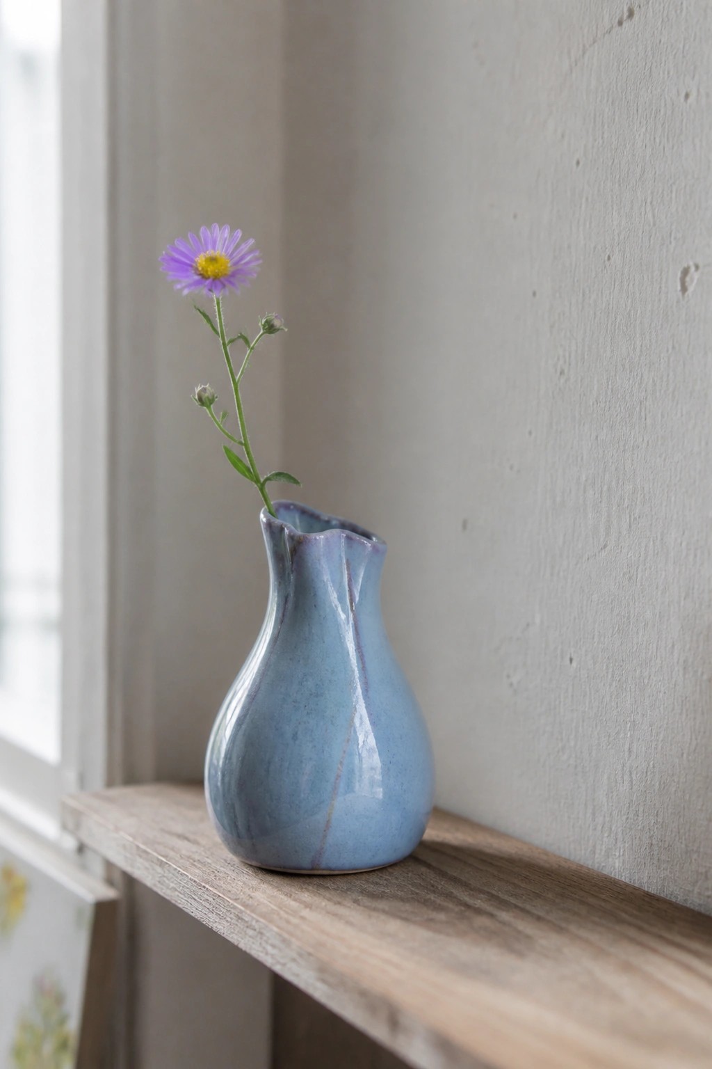 Blue ceramic vase with a purple flower on a wooden shelf against a plain wall.